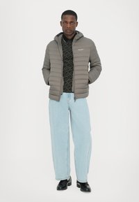 Jachetă puffer gri, matlasată, cu glugă, cu fermoar în față, purtată peste o cămașă neagră cu model, combinate cu blugi ușor albaștri și încălțăminte neagră.