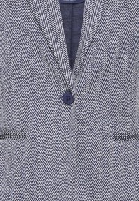 Blazer bleu marine et blanc à motif chevron, avec un seul bouton bleu marine et deux poches horizontales à l'avant.