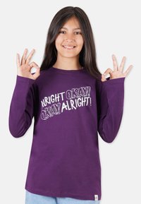 Lila langärmliges T-Shirt mit einem grafischen Schriftzug auf der Vorderseite, der "ALRIGHT OKAY! OKAY ALRIGHT!" lautet, kombiniert mit hellblauen Jeans.