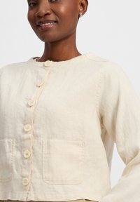 Cream-colored, linen jacket featuring a round neck, seven-button front closure, and two front pockets with slightly frayed edges.