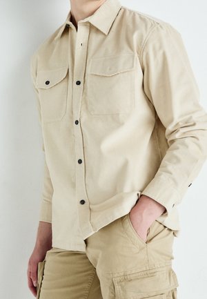 Person trägt beige kordhemd mit Knöpfen und khakifarbene Cargo-Hose, eine Hand in der Tasche, vor einfarbigem Hintergrund.
