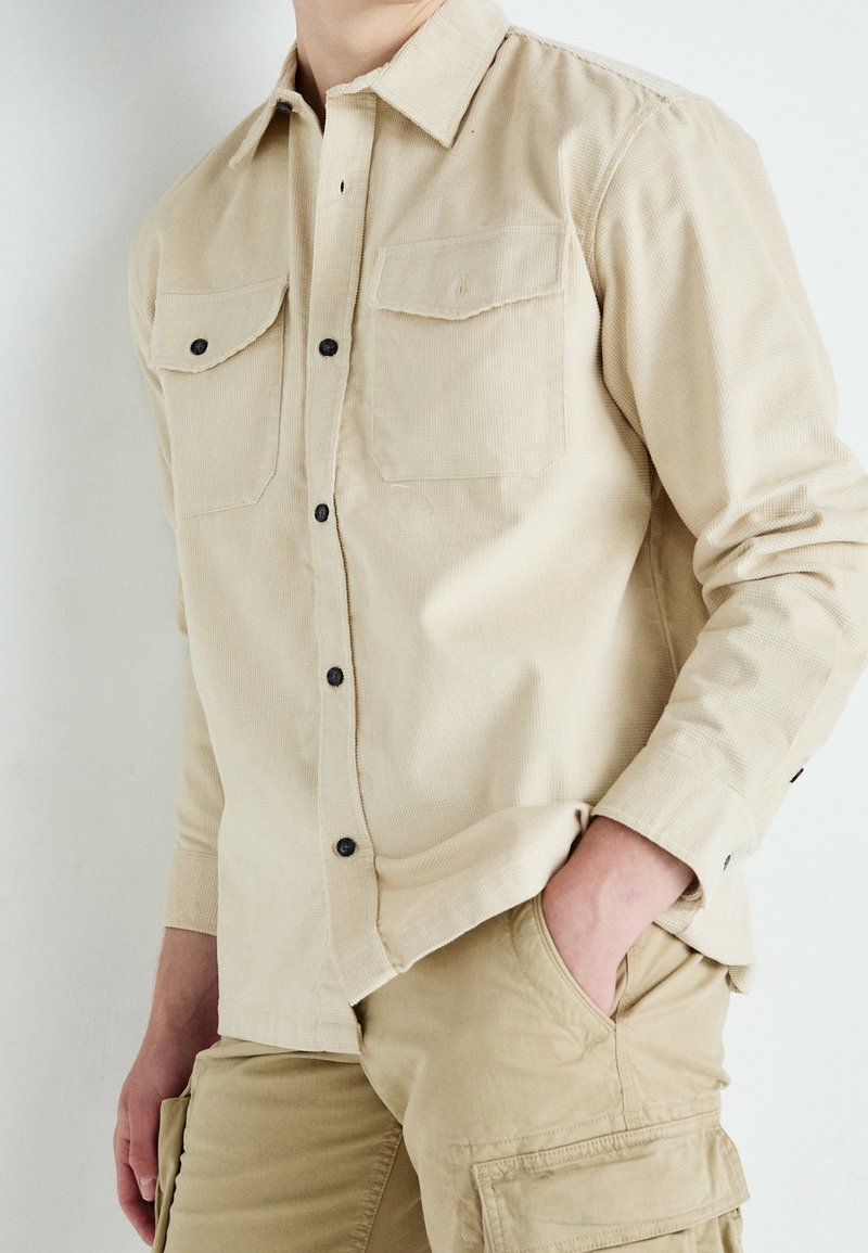 Personne portant une chemise en velours côtelé beige boutonnée et un pantalon cargo kaki, une main dans la poche, sur un fond uni.
