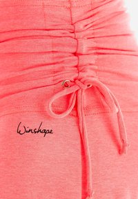 Koraļļu rozā apģērbs ar sakrautām tekstūrām, kuram ir aukla ar metāla aparatūru un melnā krāsā izšūts "Winshape" logotips.