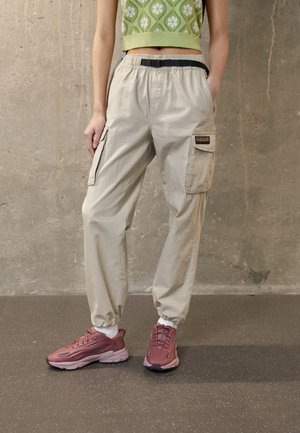 Donna che indossa pantaloni cargo beige con cintura nera, top corto verde a fiori, calzini bianchi e sneakers color malva, in piedi su pavimento scuro.