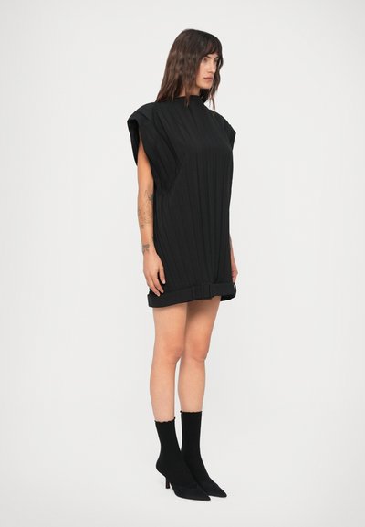 Mulher a usar um vestido preto mini canelado sem mangas com ombreiras, um detalhe de cinto na bainha, e sapatos de salto pretos pontiagudos com meias.