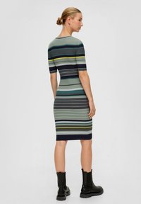 Strickkleid mit horizontalen Streifen in Grün, Marine und Gelb, figurbetont geschnitten und mit kurzen Ärmeln. Mit schwarzen Knöchelstiefeln getragen.