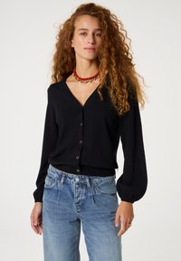 Schwarzer langer Cardigan mit V-Ausschnitt, ausgestattet mit herzförmigen Knöpfen. Kombiniert mit hellblauen High-Waist-Jeans. Glattes Material, lockere Passform.