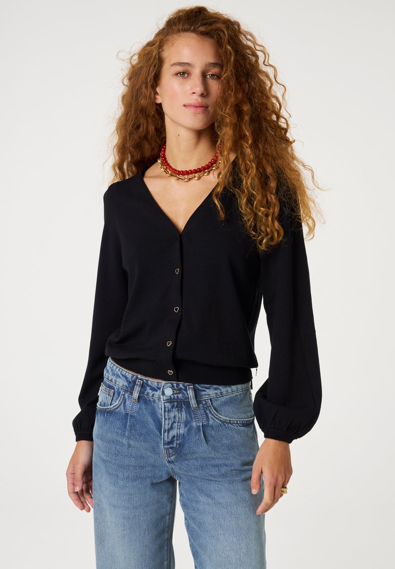 Schwarzer langer Cardigan mit V-Ausschnitt, ausgestattet mit herzförmigen Knöpfen. Kombiniert mit hellblauen High-Waist-Jeans. Glattes Material, lockere Passform.
