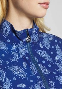 Námořnická modrá bunda s paisley vzorem v bílé barvě. Má vysoký límec, přední zip s kruhovým ozdobným táhlem a hladkou texturu.