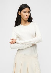 Νεαρή γυναίκα με μακριά σκούρα μαλλιά, φοράει λευκό ριμπ πλεκτό τοπ με μακριά μανίκια και ανοιχτόχρωμη πλισέ φούστα, με σταυρωμένα τα χέρια, κοιτάζοντας ελαφρώς προς τα δεξιά.