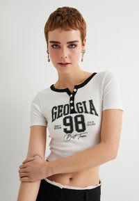 Top corto a maniche corte bianco a coste con rifiniture nere, caratterizzato da una grafica "GEORGIA 98" e dettagli con bottoni al collo, con un taglio cropped.
