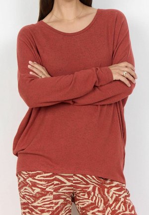 Personne portant une chemise manches longues couleur rouille, bras croisés et un pantalon à motifs avec des rayures abstraites beige et rouille.