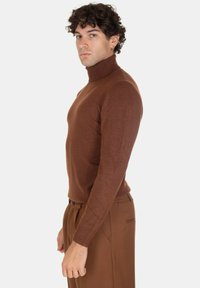 Maglione a collo alto marrone realizzato in un tessuto morbido, con un design aderente, maniche lunghe e una texture liscia. Abbinato a pantaloni marroni.
