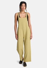 Φωτεινή χακί βαμβακερή ολόσωμη φόρμα με υφή στην επιφάνεια, φαρδιά παντελόνια, ρυθμιζόμενους ιμάντες και δύο μπροστινές τσέπες. Συνδυασμένη με μαύρο τοπ και ξύλινα παπούτσια clogs.