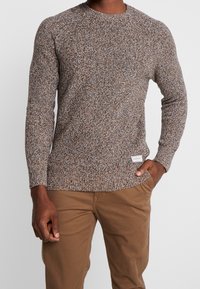 Brun texturerad stickad tröja med rund halsringning och långa ärmar, som har en liten varumärkesetikett på den nedre sidan. Matcher med ljusbruna byxor.