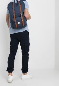 Granatowy plecak z brązowymi skórzanymi paskami, prostokątny kształt, materiał tekstylny, regulowane paski oraz przednia kieszeń; noszony z spodniami cargo i trampkami.