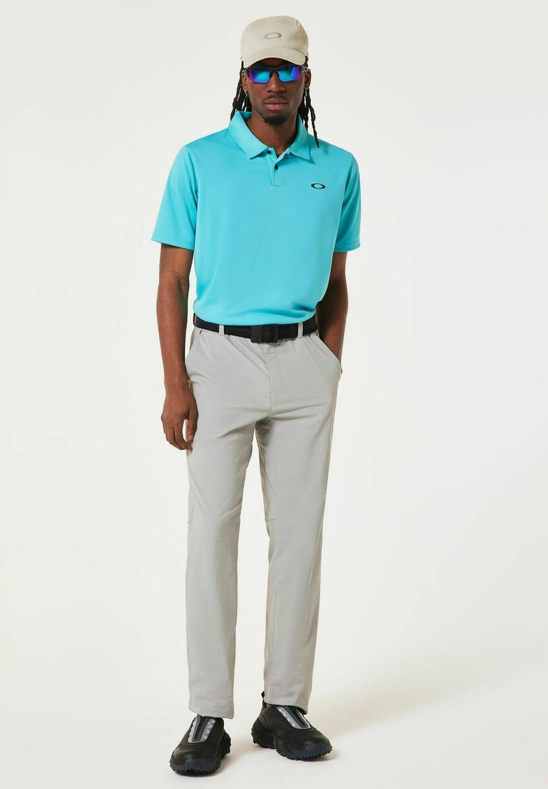 Polo turquoise, pantalon gris clair, ceinture noire, chaussures noires à semelles texturées, casquette beige, lunettes de soleil bleues, style décontracté.