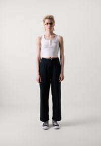 Bílý zkrácený top bez rukávů s henley výstřihem, kombinovaný s tmavými širokými kalhotami a černo-bílými teniskami. Obsahuje sluneční brýle a piercingy v uších.
