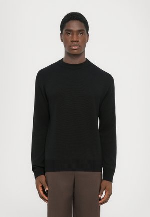Schwarzer Rundhals-Pullover mit langen Ärmeln, strukturiertem Strickstoff und geripptem Saum. Kombiniert mit braunen Hosen.