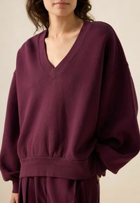 Sweatshirt oversize bourgogne avec un col en V, un ourlet côtelé et des manches larges en tissu doux, mettant en avant une coupe décontractée et une texture lisse.