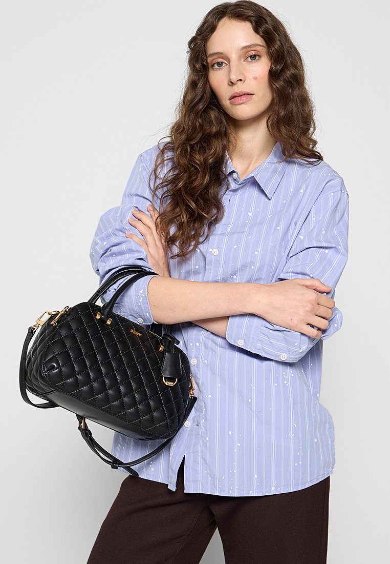 Gepolsterte schwarze Handtasche mit goldenem Zubehör, ausgestattet mit zwei Obergriffen und einem abnehmbaren Schulterriemen. Getragen mit einer hellblauen gestreiften Bluse.