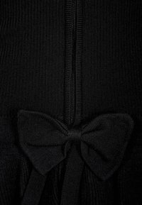 Lähedalt vaade mustale ribakujulisele kangale, millel on keskel tõmblukk ja alumises osas kinni seotud kudumislint.