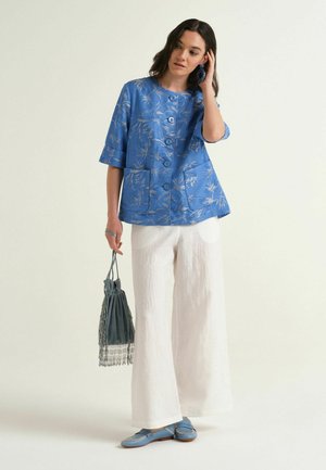 Donna che indossa una giacca blu floreale con bottoni, pantaloni bianchi a gamba larga, mocassini blu, tiene una borsa grigia con frange e si tocca i capelli.