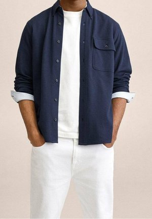 Homme portant une chemise bleu marine boutonnée sur un t-shirt blanc avec un pantalon blanc, mains dans les poches, manches de chemise retroussées.