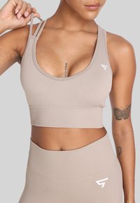 Beżowy stanik sportowy z okrągłym dekoltem i wieloma cienkimi ramiączkami. Posiada żebrowany dół oraz akcent z logo po lewej stronie. Gładka tekstura.