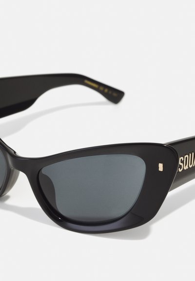 Black sunglasses with angular cat-eye shape, dark lenses, glossy frame, and gold hardware on the temple featuring the brand name.