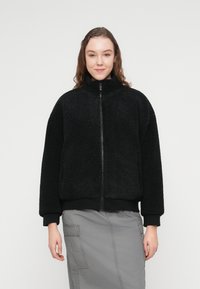 Μαύρο ζακέτα από φλις με ψηλό γιακά και φερμουάρ, με υφή στην επιφάνεια και ριμπ μανσέτες. Φορεμένο πάνω από μια γκρι, μέσης μήκους φούστα.