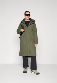 Palton lung de culoare verde olive, impermeabil, cu glugă, fermoar frontal și șnururi ajustabile. Combinat cu pantaloni negri și încălțăminte de drumeție gri.