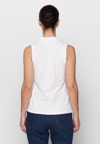 Bílý bezrukávový polo top s límečkem, který má hladkou strukturu a přiléhavý střih, spárovaný s tmavě modrými džíny.