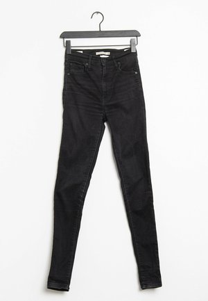 Vaqueros pitillo negros hechos de mezclilla, con cintura de talle alto, cinco bolsillos y una textura suave. Colgados en una percha negra.