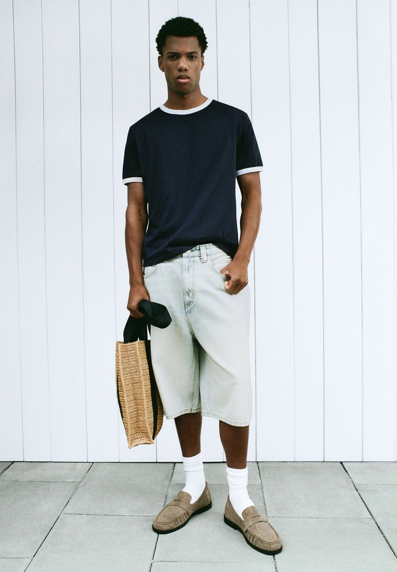 Un homme se tient contre un mur à panneaux blancs, portant un t-shirt bleu marine, un short en jean clair jusqu'aux genoux, des chaussettes blanches, des mocassins marron et tenant un sac cabas tressé.