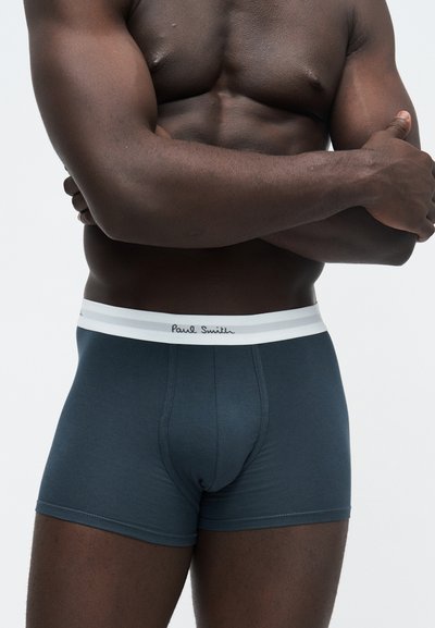 Μυώδης άνδρας με σκούρο δέρμα που φοράει σκούρα μπλε μποξεράκια Paul Smith με λευκή μέση, τα χέρια σταυρωμένα πάνω στο γυμνό στήθος.