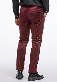 Bordó bársonynadrág, slim fit fazonnal, fekete oldalsávokkal, valamint két hátsó zsebbel, gombos záródással. Fekete cipők láthatók.