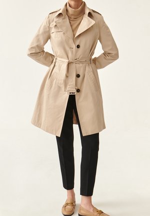 Femme portant un trench-coat beige ceinturé sur un col roulé couleur tabac, pantalon slim noir et mocassins beiges, debout les mains dans le dos.