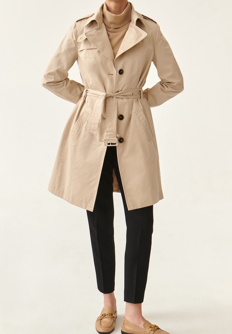 Femme portant un trench-coat beige ceinturé sur un col roulé couleur tabac, pantalon slim noir et mocassins beiges, debout les mains dans le dos.