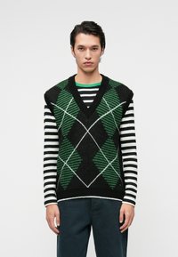 Schwarze Weste mit grünem Rauten- und Streifenmuster, mit V-Ausschnitt. Getragen über einem schwarz-weißen gestreiften Langarmshirt.