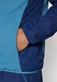 Windbreaker in Blau und tiefem Marineblau; verfügt über eine glatte Textur und elastische Bündchen; enthält eine Seitentasche mit einem verdeckten Öffnung.