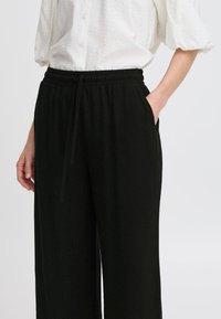Schwarze weit geschnittene Hose mit elastischem Bund und Kordelzug. Taschen auf jeder Seite. Der Stoff wirkt weich mit einer glatten Textur.