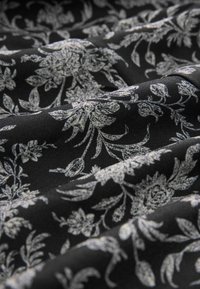 Černá látka s jemným bílým květinovým vzorem. Hladká textura s propletenými listy a květy vytvářejícími decentní, elegantní design.