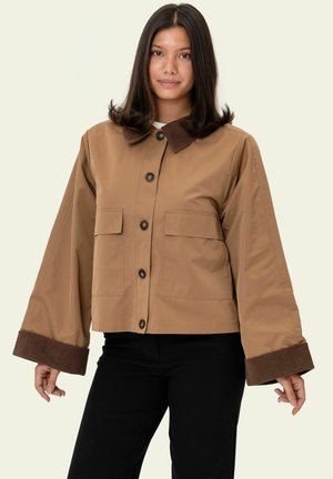 Naine, kandes heledat nööbitavat jopet tumepruuni kraa ja mansettidega ning musti pükse, seisab lihtsa heleda taustaga ees.