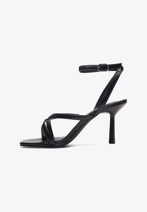 Sandale à talon noir avec des brides croisées, un bout carré et une bride de cheville avec une boucle. Fabriquée en cuir lisse avec une finition mate.