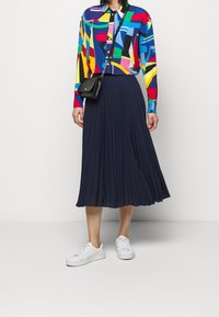 Kolorowa koszula z wzorami w geometrie, zestawiona z plisowaną granatową spódnicą i białymi trampkami, nosząc małą czarną torbę.