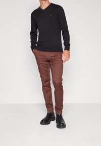 Maglione nero a V abbinato a pantaloni slim-fit marroni e stivaletti neri. Il maglione presenta un piccolo logo sul petto.