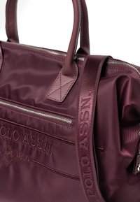 Viininpunainen nailonlaukku nahkakoristein, vetoketjusuljenta ja etutasku. Varustettu kirjotulla logolla ja säädettävällä kangashihnalla olkapäälle.