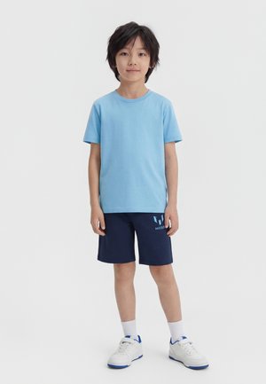 Chlapec s čiernymi vlasmi, oblečený v svetlomodrom tričku, tmavomodrých šortkách s logom, bielych ponožkách a bielych teniskách, stojaci pred jednofarebným pozadím.