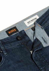 Mörkblå denimjeans med dragkedja och knappstängning. Etiketter inkluderar "BOSS" och "SOFT MOTION." Stickning är orange.